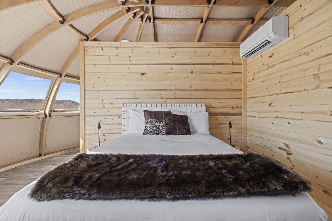 Alt text: "Interior of a geodesic dome house featuring a cozy bedroom with wooden walls, a comfortable bed, and large porthole windows offering scenic views. Ideal for luxury glamping or a stylish backyard office. Keywords: geodesic dome, dome home, glamping pods."