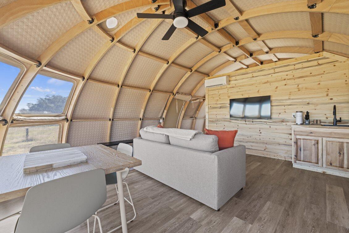 Alt text: "Interior of a stylish geodesic dome home featuring wooden paneling, a cozy seating area, and large porthole windows. Ideal for glamping, sustainable living, or as a luxury Airbnb option. The space includes a modern kitchenette and a dining area, showcasing the versatility of geodesic dome kits for creating unique living spaces."
