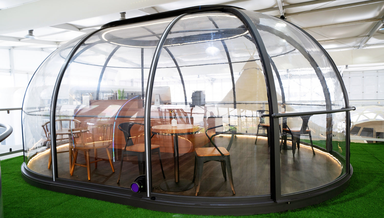 Alt text: "Modern geodesic dome with transparent walls, featuring a stylish interior setup with wooden chairs and a table, ideal for glamping or as a backyard office. This dome structure exemplifies luxury and versatility, perfect for sustainable living or creating a unique guest space. Keywords: geodesic dome, glamping pod, artemis backyard office."