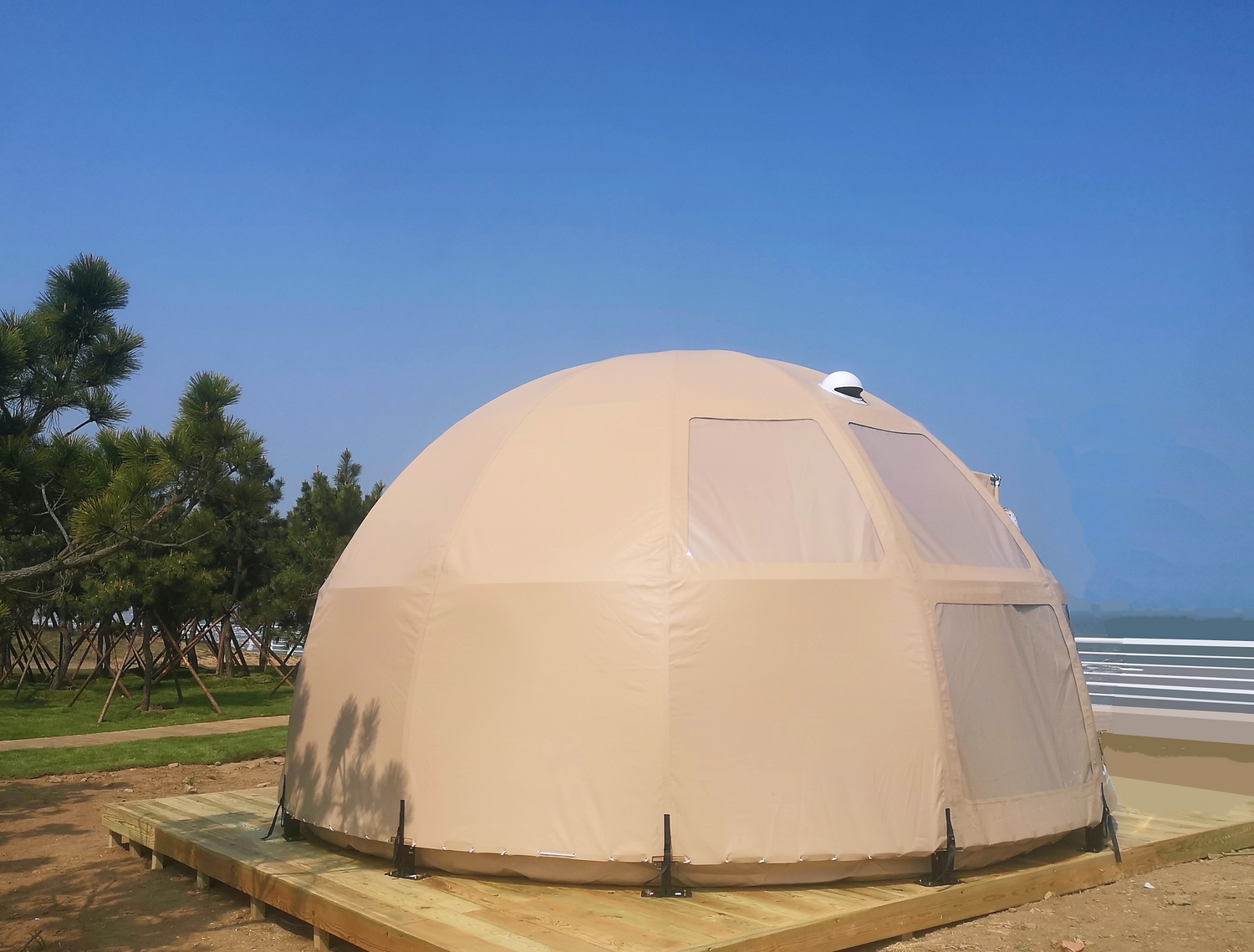 Alt text: "Large beige geodesic dome tent with porthole windows set on a wooden platform, surrounded by trees, ideal for glamping or as a backyard dome office. High-quality, stylish, and cost-effective solution for sustainable living or luxury Airbnb experiences."
