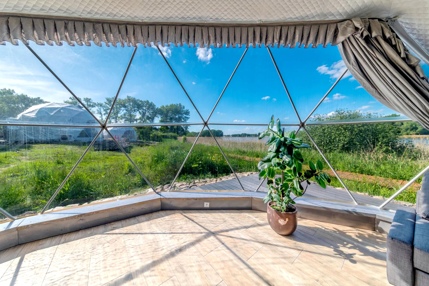 Alt text: "Interior view of a geodesic dome with large porthole windows, showcasing a scenic outdoor landscape. The space features a potted plant and stylish furnishings, ideal for glamping or use as a luxury Airbnb. This dome home offers a sustainable and cost-effective solution for those seeking a unique living or recreational space."