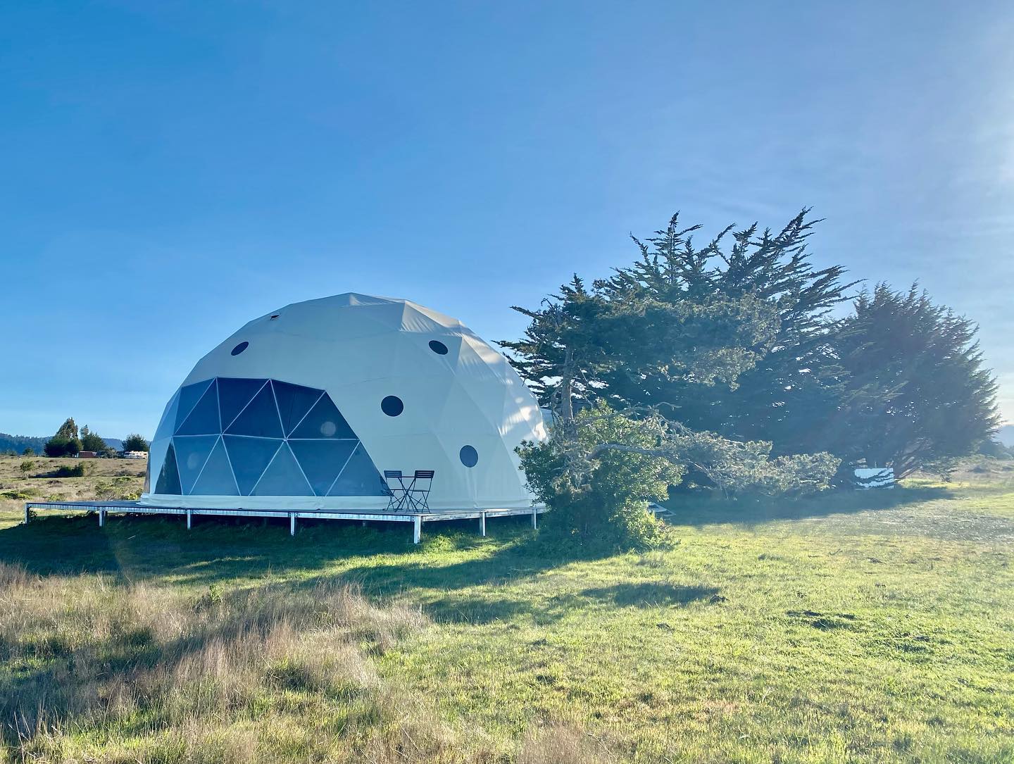 A large geodesic dome with porthole windows set in a scenic outdoor location, ideal for glamping or use as a luxury Airbnb. The dome features a spacious design, perfect for sustainable living or as a backyard office.