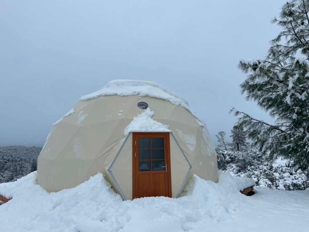 Alt text: "Snow-covered geodesic dome with wooden door and porthole window, ideal for glamping or sustainable living. Perfect as a luxury Airbnb or backyard office, showcasing the versatility of geodesic dome house kits."