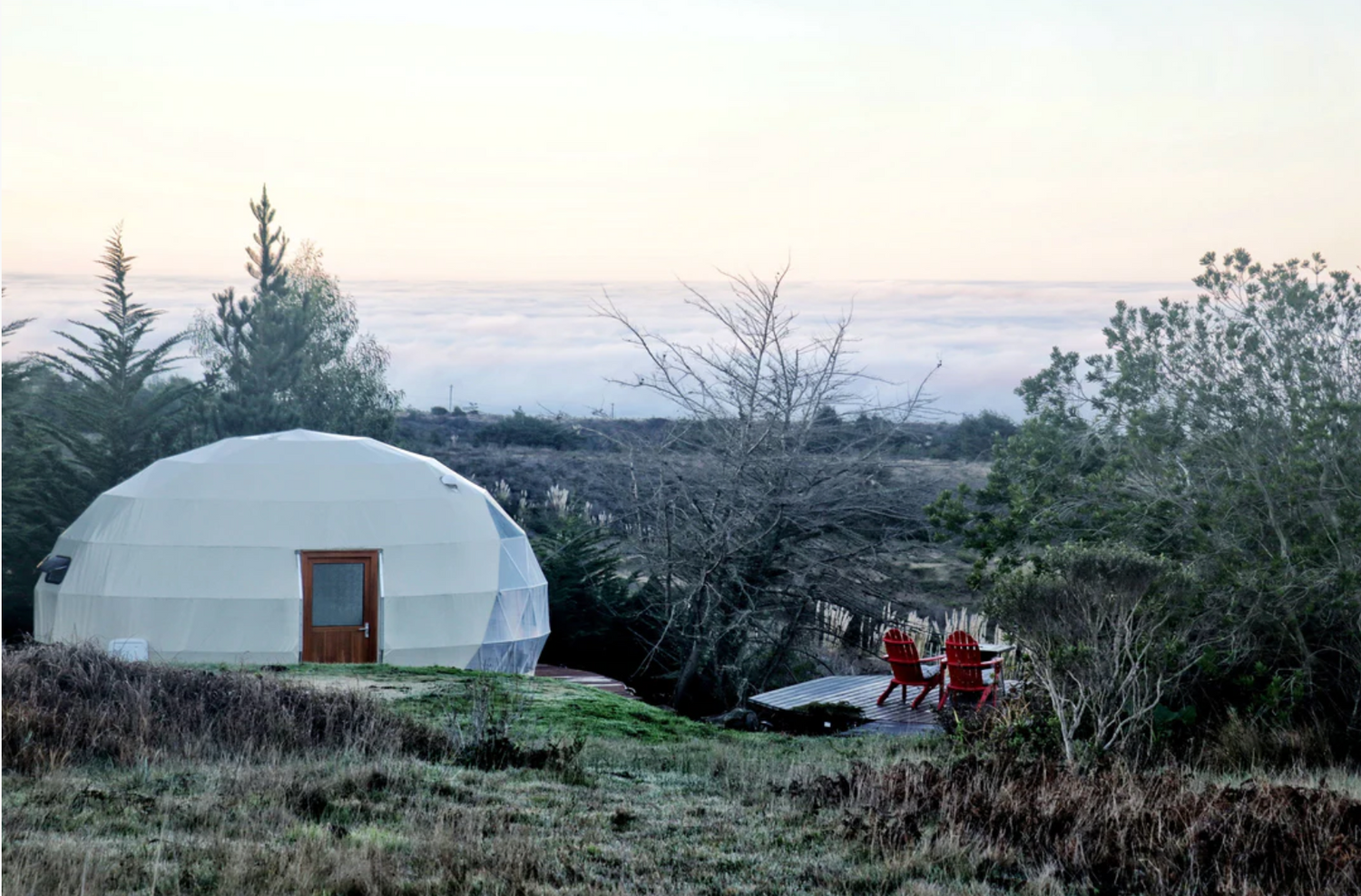Alt text: "Geodesic dome house with porthole windows set in a scenic outdoor landscape, featuring red chairs on a wooden deck. Ideal for glamping, sustainable living, or as a luxury Airbnb option. High-quality dome home solution by a company specializing in glamping pods and geodesic dome kits."