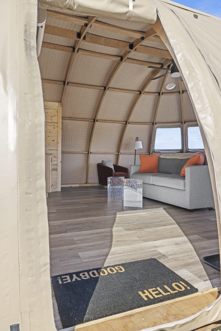 Alt text: &quot;Interior view of a stylish geodesic dome with wooden framework, featuring a cozy seating area with a gray sofa and orange cushions, a modern lamp, and porthole windows. Ideal for glamping, backyard offices, or sustainable living. Keywords: geodesic dome, glamping pod, dome home, luxury Airbnb.&quot;