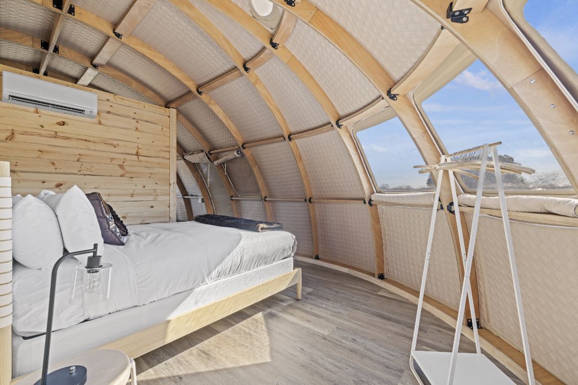 Interior view of a stylish geodesic dome with wooden accents, featuring a cozy bed and large porthole windows, ideal for luxury glamping or a unique backyard office.