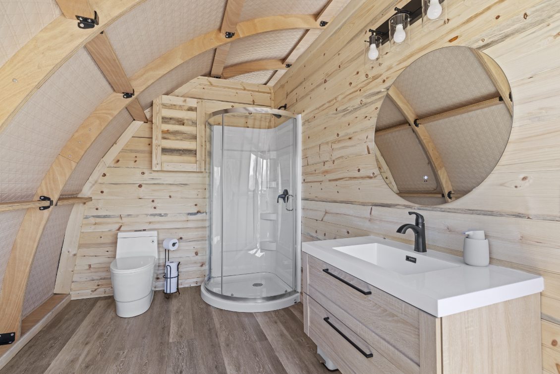 Alt text: &quot;Interior of a stylish geodesic dome bathroom featuring wooden walls, a modern shower, toilet, and vanity with a round mirror. Ideal for luxury glamping or a geodome home setup.&quot;