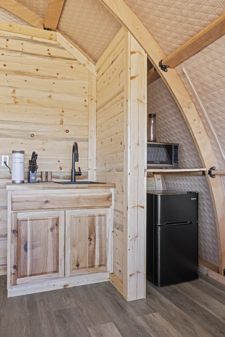 Alt text: &quot;Interior view of a wooden glamping pod featuring a kitchenette with a sink, cabinets, and a small refrigerator. The stylish design highlights the use of natural wood, ideal for sustainable living or a luxury Airbnb experience. Perfect for those interested in geodesic dome house kits or backyard dome solutions.&quot;