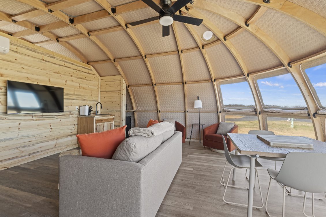 Interior view of a stylish geodesic dome home featuring a cozy living area with a sofa, chairs, and a dining table. The space is well-lit with natural light streaming through large porthole windows, showcasing the scenic outdoor landscape. This luxury glamping pod offers a modern and sustainable living solution, ideal for those seeking a unique and comfortable experience in a dome house.