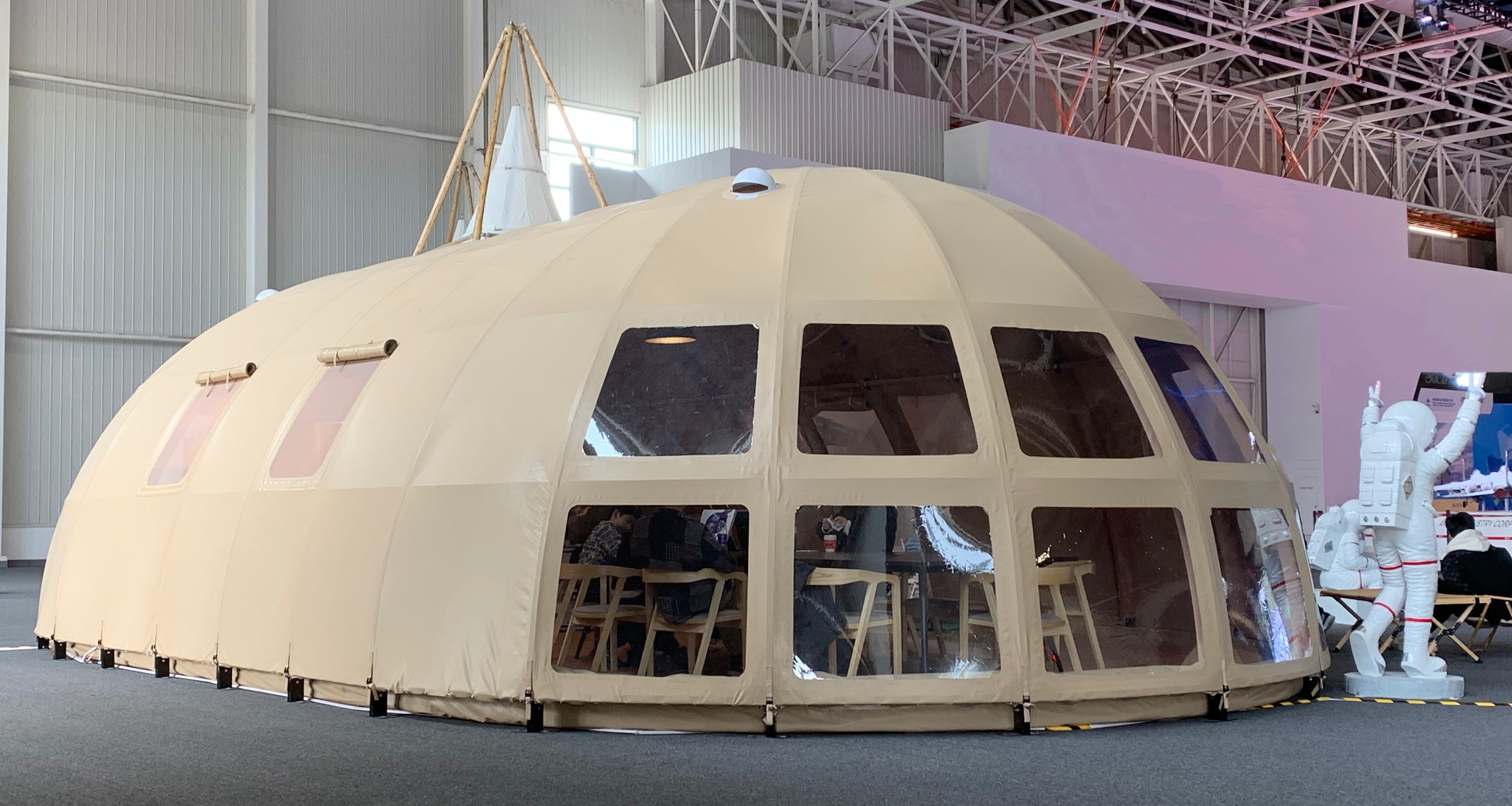 A large beige geodesic dome tent with clear porthole windows, set up indoors. The dome is designed for versatile use, such as glamping, sustainable living, or as a backyard office. Inside, wooden chairs are visible, highlighting its spacious interior. An astronaut figure stands nearby, adding a unique touch to the display.