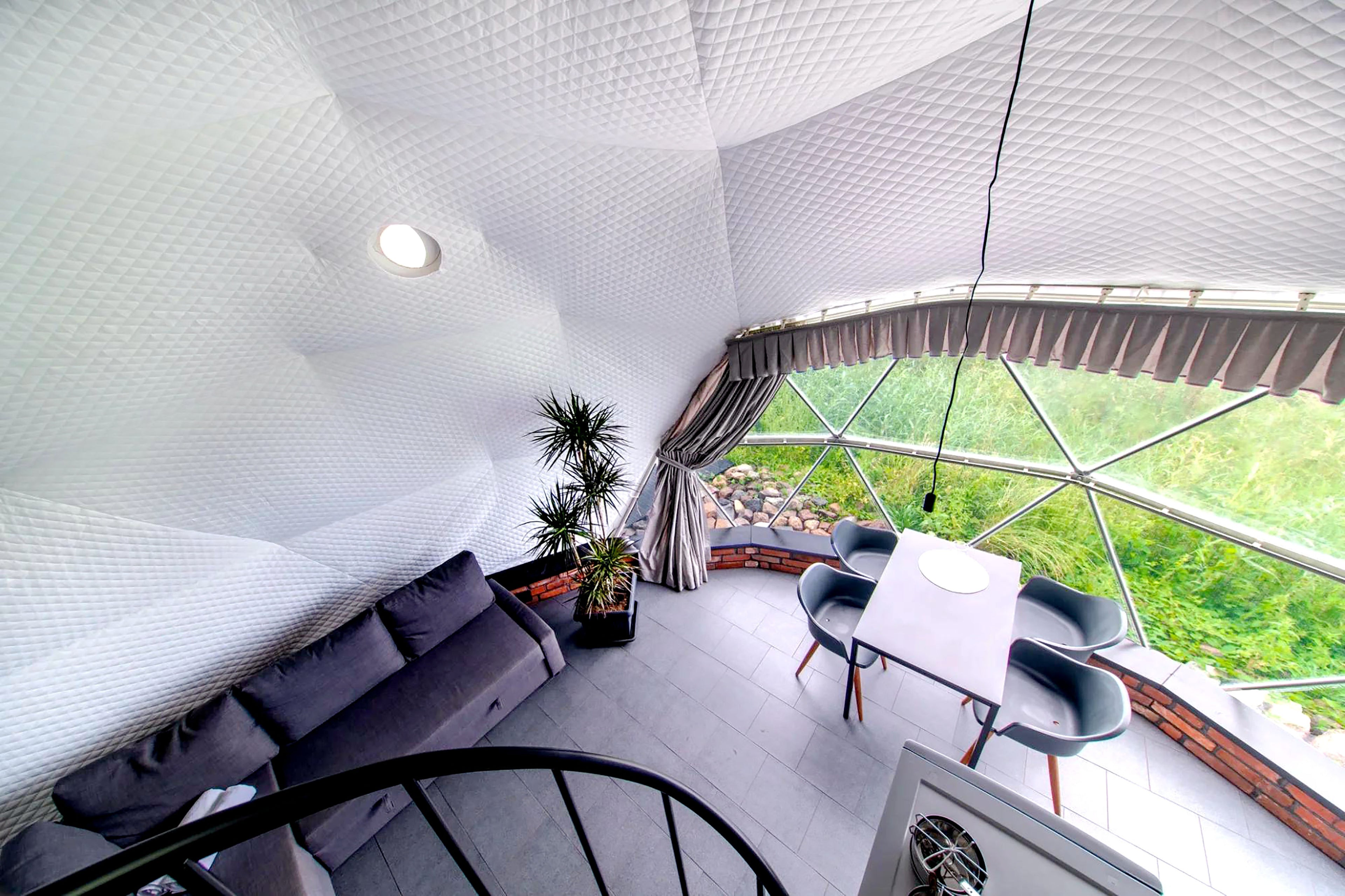 Interior view of a stylish geodesic dome with a modern setup, featuring a cozy seating area, dining table, and large porthole windows offering a scenic outdoor view. Ideal for luxury glamping, sustainable living, or as a unique backyard office space.