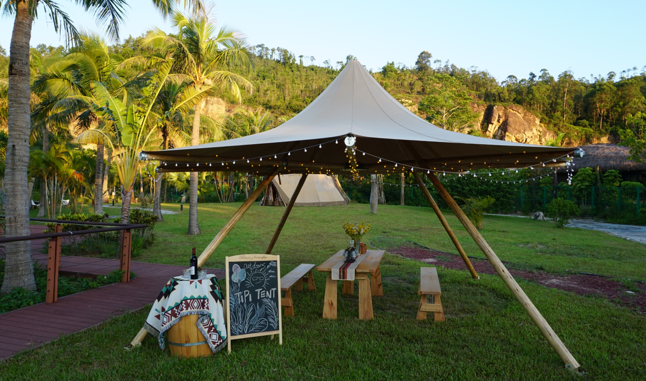 Outdoor glamping setup featuring a stylish tipi tent with string lights, surrounded by lush greenery. A wooden picnic table and a decorative barrel with a chalkboard sign enhance the inviting atmosphere, ideal for luxury camping experiences. Keywords: glamping, tipi tent, outdoor dome, luxury Airbnb.