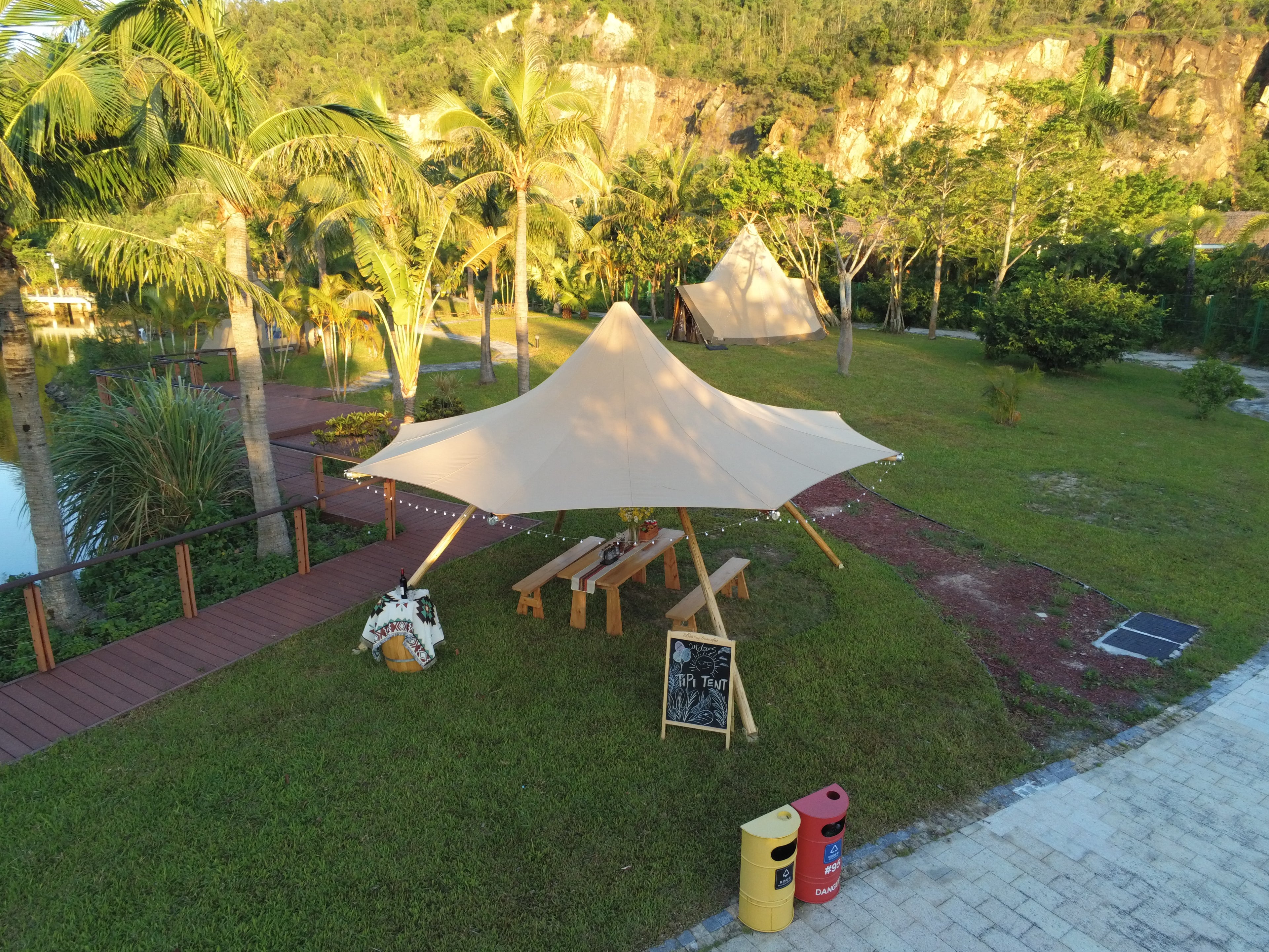 A scenic outdoor setting featuring a stylish glamping setup with a large bell tent and picnic area, surrounded by lush greenery and palm trees. Ideal for those seeking luxury camping experiences or sustainable living solutions. Keywords: glamping, bell tent, outdoor dome, luxury camping, geodesic dome.