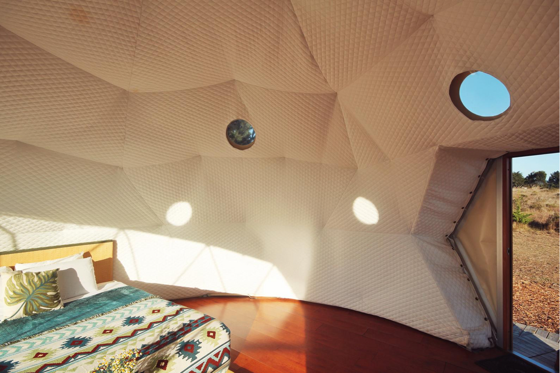 Interior view of a stylish geodesic dome with porthole windows, featuring a cozy bed with patterned bedding. Ideal for luxury glamping or as a unique backyard office space.