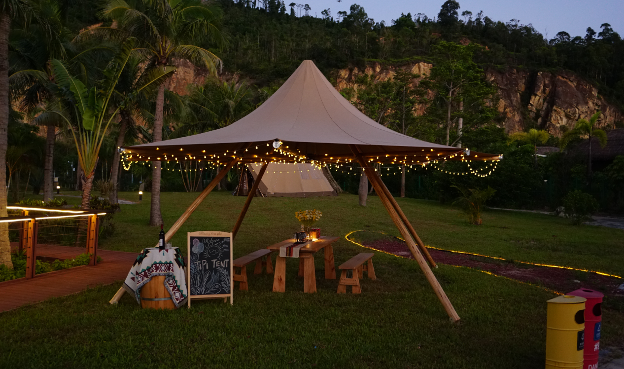 Outdoor glamping setup featuring a stylish teepee tent adorned with string lights, surrounded by lush greenery. A wooden picnic table with benches is set underneath, creating a cozy atmosphere for relaxation. Ideal for those seeking luxury camping experiences or unique backyard retreats.