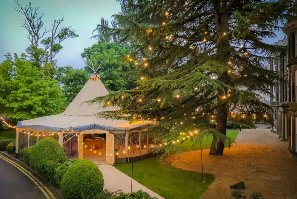 Alt text: &quot;Elegant glamping setup featuring a large bell tent illuminated with string lights, surrounded by lush greenery. Ideal for luxury outdoor experiences, this stylish tent offers a premium glamping solution for those seeking a unique and comfortable stay.&quot;