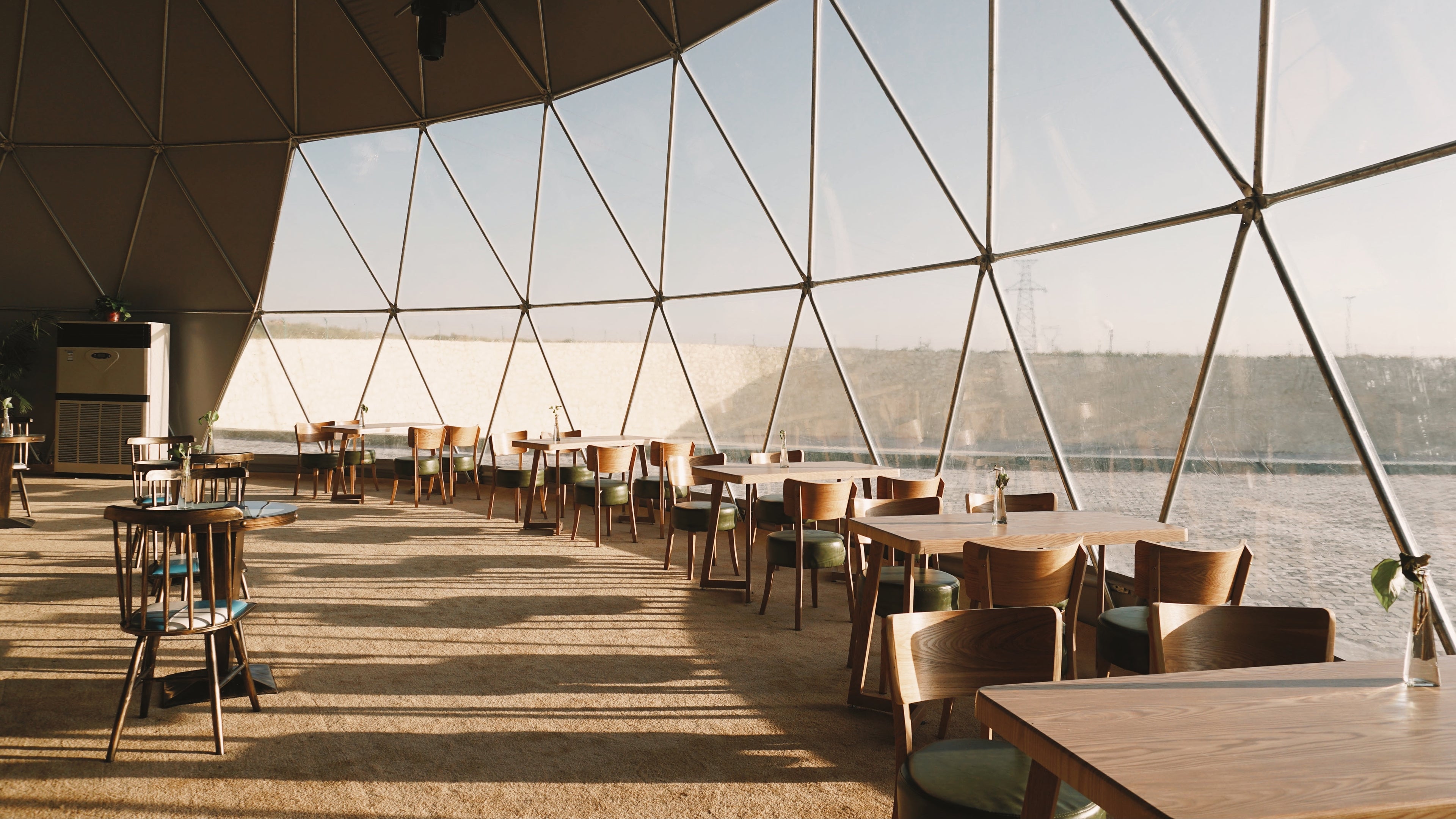 Interior view of a spacious geodesic dome with wooden tables and chairs, showcasing a stylish and modern design. The large windows provide ample natural light, ideal for a luxury Airbnb or a unique glamping experience. Perfect for those interested in geodesic dome house kits or creating a sustainable living space.