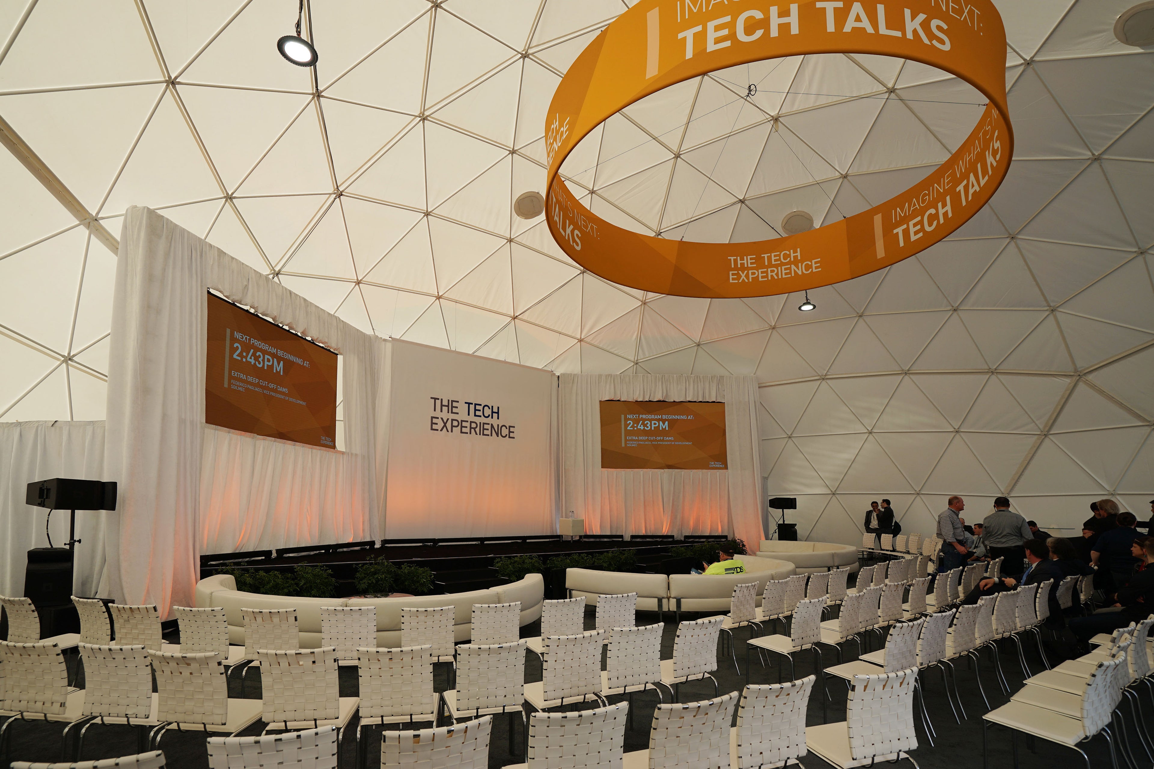 A spacious geodesic dome set up for a tech event, featuring rows of white chairs and a stage with &quot;The Tech Experience&quot; branding. The dome&