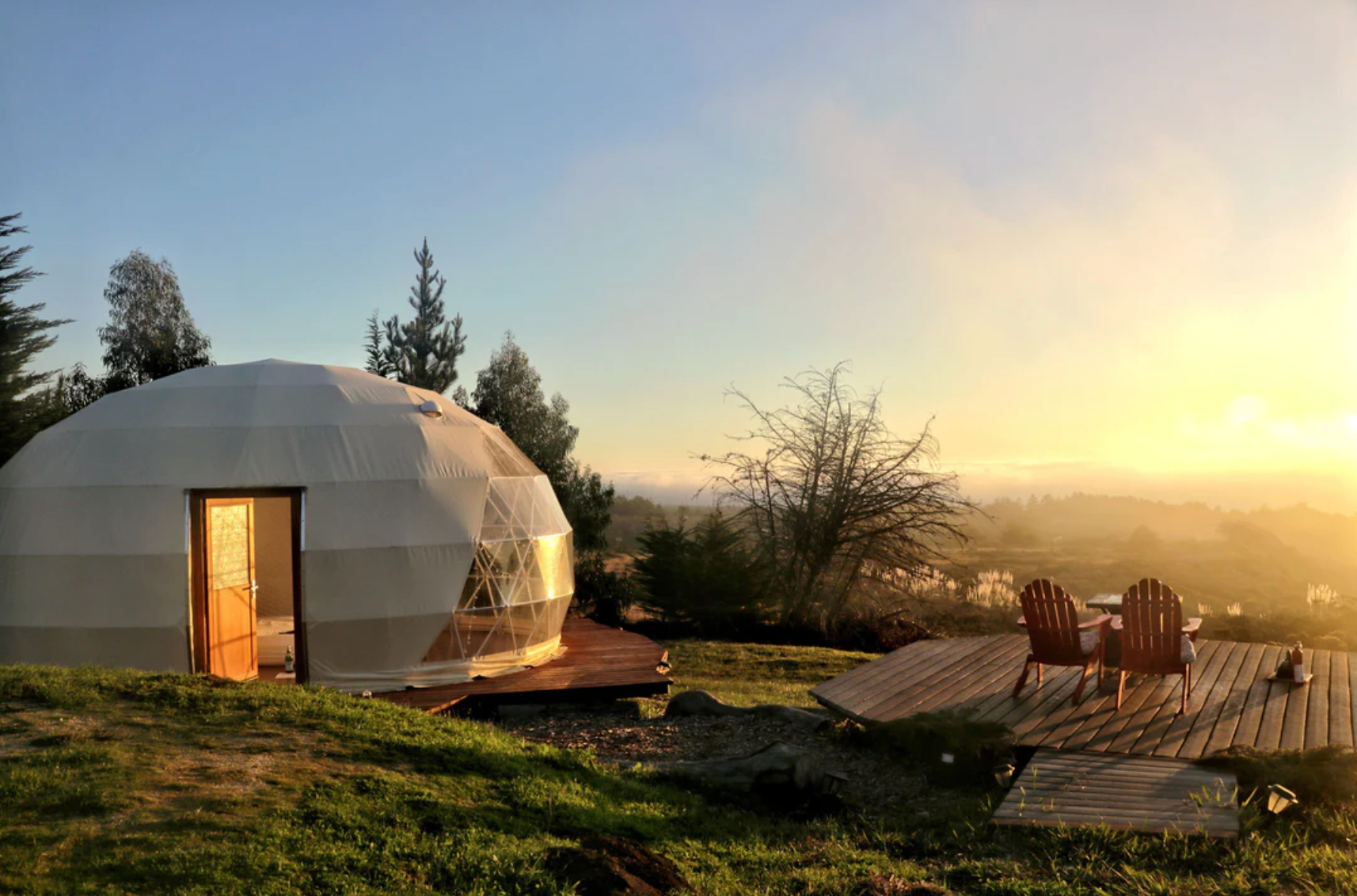 Alt text: &quot;Scenic view of a geodesic dome house with porthole windows at sunset, surrounded by nature. The dome, ideal for glamping or as a backyard office, is set on a wooden deck with two chairs, offering a luxurious and sustainable living solution.&quot;