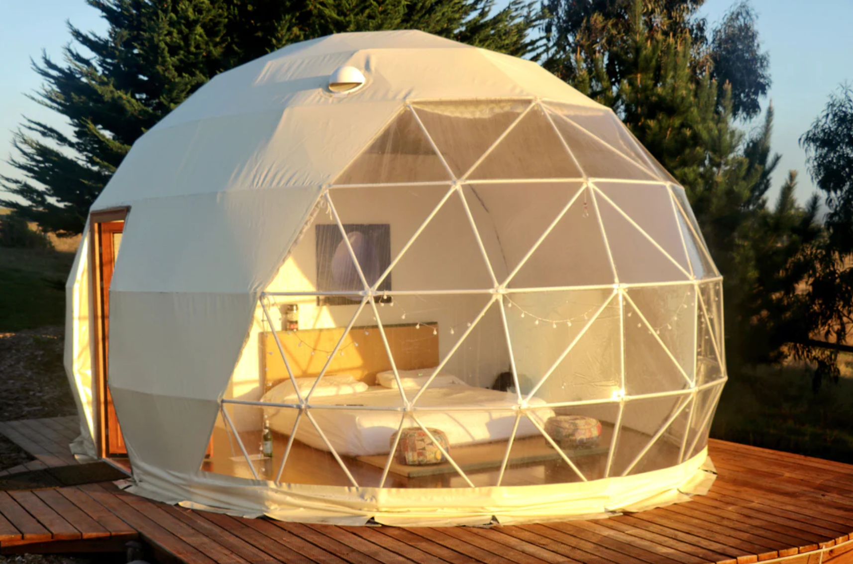 Alt text: &quot;Luxury geodesic dome with porthole windows set up for glamping, featuring a cozy interior with a bed, located on a wooden deck surrounded by trees. Ideal for sustainable living, art studios, or as a stylish backyard dome. Available from a company specializing in high-quality glamping solutions.&quot;