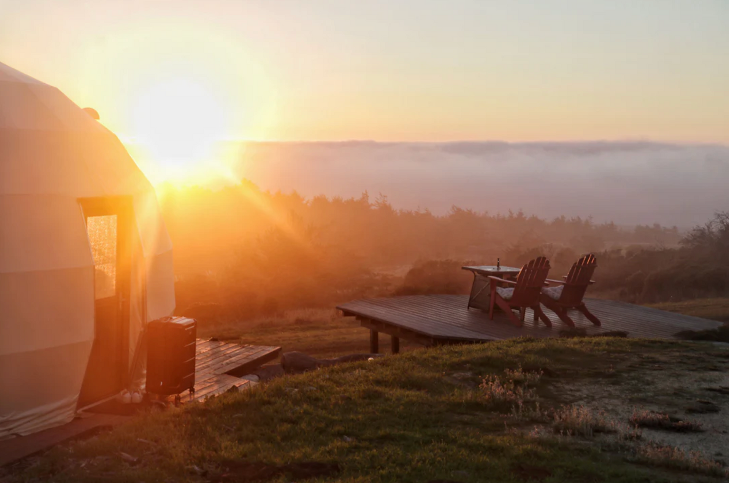 Sunset view of a luxurious geodesic dome with a wooden deck and chairs, ideal for glamping or a backyard office. The dome, featuring porthole windows, is set in a serene landscape, showcasing the company&