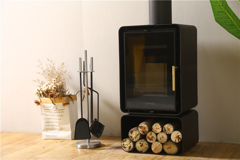 A modern black wood-burning stove with a glass door, accompanied by a set of fireplace tools and a decorative arrangement of dried flowers in a paper bag. Logs are neatly stacked in a compartment below the stove, set against a light-colored wall and wooden floor. Perfect for enhancing the cozy atmosphere in a geodesic dome house or glamping pod.