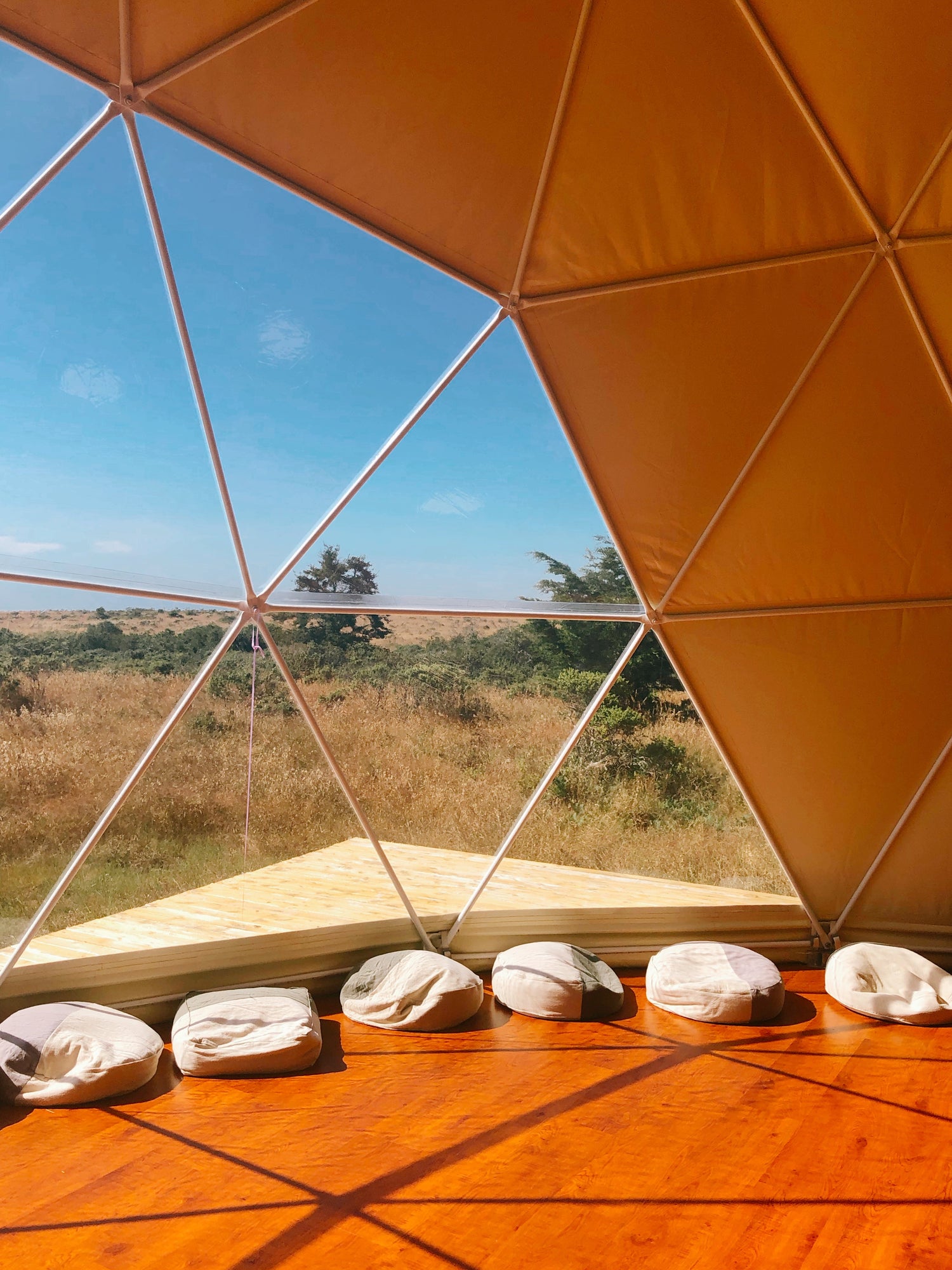 Interior view of a geodesic dome with large porthole windows, showcasing a wooden floor and round cushions. This spacious dome is ideal for glamping, offering a luxurious and sustainable living space. Perfect for those interested in geodesic dome house kits, backyard domes, or creating a unique outdoor retreat.