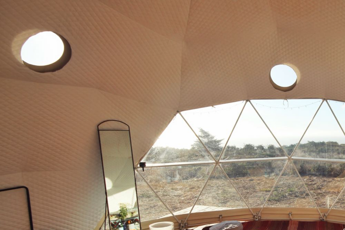 Interior view of a geodesic dome with porthole windows, showcasing a stylish and spacious design ideal for glamping or a luxury Airbnb. The dome features a large window offering a scenic outdoor view, perfect for sustainable living or creating a unique backyard office.