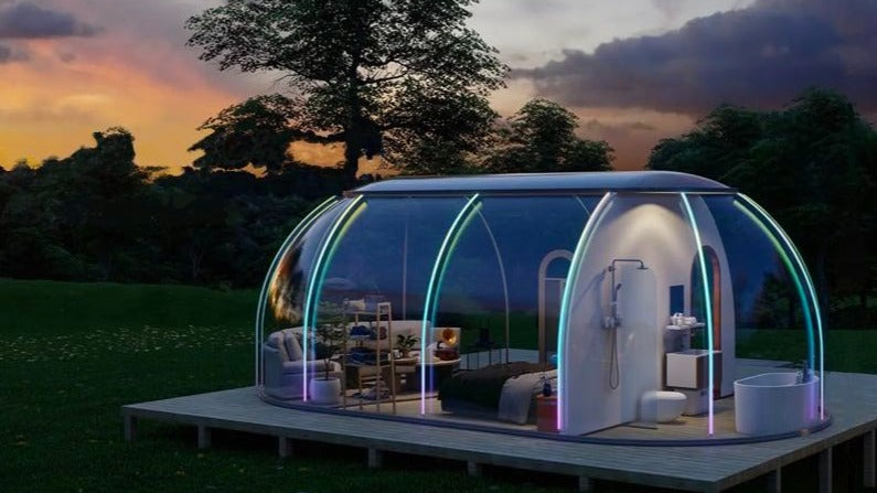 A modern, transparent geodesic dome with porthole windows set in a scenic outdoor environment at dusk, showcasing a stylish interior with a bed and bathroom, ideal for luxury glamping or a unique backyard office.