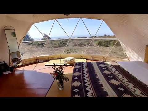 Interior view of a stylish geodesic dome with large porthole windows, featuring a cozy bed and scenic outdoor landscape. Ideal for luxury glamping or as a unique backyard office space.